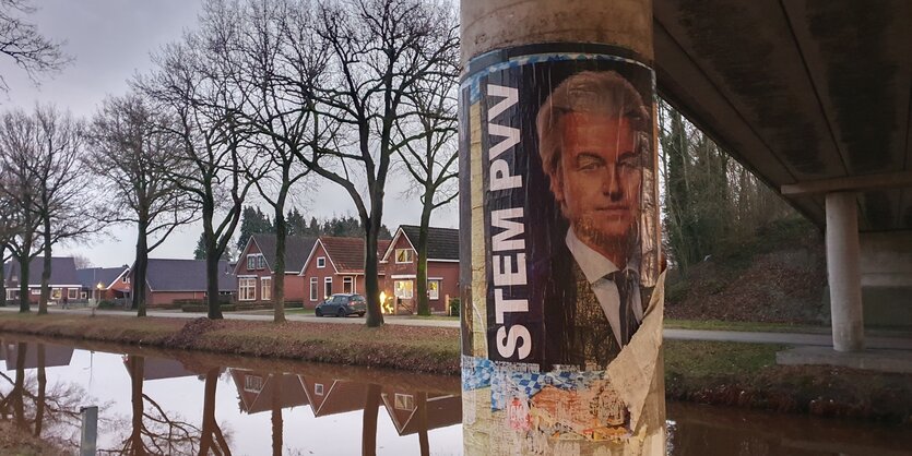 An einer Brückensäule klebt ein beschädigtes Wahlplakat von Geert Wilders, im Hintergrund sind ein gut gefüllter Kanal, Bäume und rote Klinkerhäuser zu sehen.