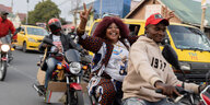 Eine Frau auf dem Beifahrersitz eines Motorrads trägt ein T-shirt, auf das das Konterfei des kongolesischen Präsidenten Tshisekedi gerduckt ist und macht das Victory-Zeichen mit ihren Händen