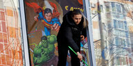 Eine Frau räumt Schutt aus einem zerbombten Fenster, im Hintergrund sieht man Superman und Hulk an der Wand des Zimmers gemalt