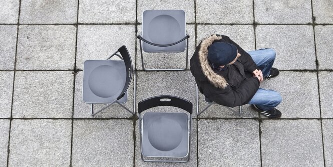 Vier Stühle stehen in einem Stuhlkreis, die Rückenlehnen sind einander zugewandt, auf einem Stuhl sitzt ein mann