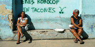 Zwei Frauen sitzen auf einem Hausvorsprung in der Sonne. Zwischen ihnen steht an der türkisen Hauswand "Tu eres Robocop en tacones".