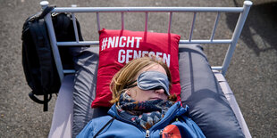 Protest im öffentlichen Raum - eine Frau liegt mit Schlafmaske auf einem Metallbett. Auf dem roten Kissen steht #nicht genesen