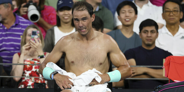 Tennisprofi Rafael Nadal sitzt mit nackten Oberkörper und angestrengtem Gesichtsausdruck auf der Bank, hinter ihm die Match-Zuschauer