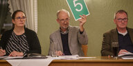 Drei Menschen am Tisch, der mittlere hebt ein Blatt mit der Nummer "5" hoch