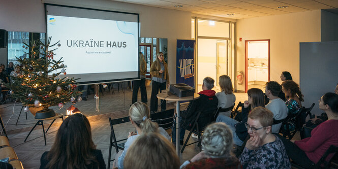 Eine Frau hält eine Präsentation, auf Stühlen sitzen mehrere Personen und hören zu