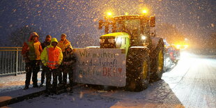 Fünf Menschen in Warnwesten stehen im Schneegestöber nachts neben einem Traktor, an dem ein Transparenz befestigt ist: "Wir machen Landwirtschaft auch für Dich!"