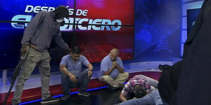 Ein maskierter Mann mit einem Gewehr steht in einem Fernsehstudio, mehrere Menschen liegen am Boden