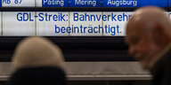 Bahnreisende stehen vor einer Anzeigetafel mit der Aufschrift „GDL-Streik: Bahnverkehr beeinträchtigt.“.