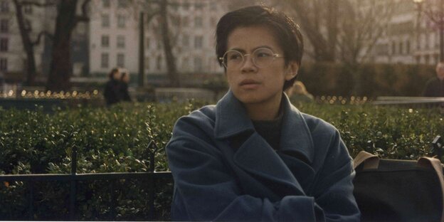 Qiu Miaojin sitzt auf einer Bank , dahinter eine Grünfläche, sie trägt einen taubenblauen Wintermantel und schaut zur Seite