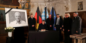 Markus Söder sitzt vor einem Bild von Franz Beckenbauer und schreibt. Verschiedene Personen stehen um ihn herum.