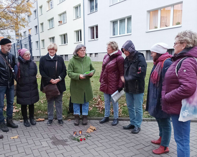 Ein Gruppe ältere Menschen steht um einen Stolperstein herum, neben der Gedenktafel stehen Grabeskerzen.