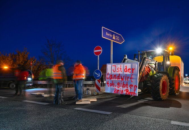 Ein Traktor mit Transparent steht auf eine Autobahnauffahrt, davor Menschen mit Warnwesten