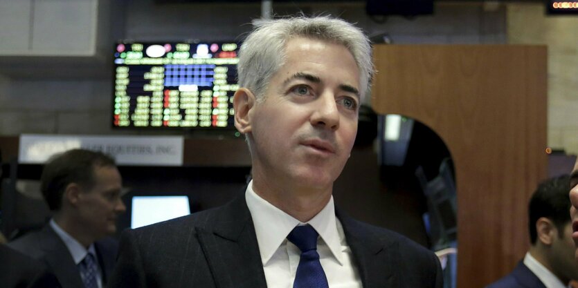 Bill Ackman, ein älterer Mann mit kurzen grauen Haaren. Er trägt einen dunklen Anzug und eine Dunkle Krawatte. Hinter ihm ein Monitor mit verschwommenen tabellen, der von der Decke hängt. Mehrere Männer laufen im Hintergrund durchs Bild. Sie tragen auch Anzug.