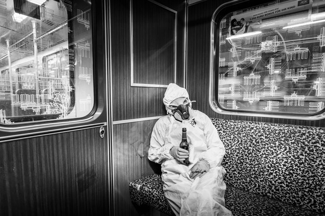 Person in Schutzanzug und mit Bierflasche in der Hand sitzt in der U-Bahn