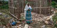 Eine ältere Frau in bunter Kleidung steht auf einen Stock gestützt vor ihrem zerstörten Haus