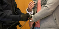 Einem Mann in gemusterter Strickjacke werden die Handschallen von einem Justizbeamten abgenommen