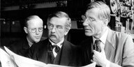 Auf einem Schwarz-Weiß-Filmstill lesen drei Männer im Anzug eine Zeitung