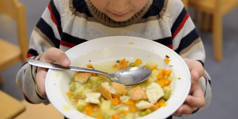 Ein Kind hält einen Teller mit Gemüsesuppe in der Hand.