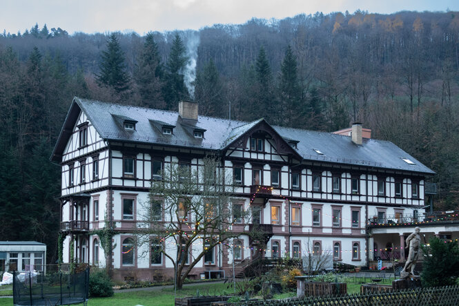 Rauch steigt aus einem sehr großen alten Haus, dahinter Wald vor Bergen