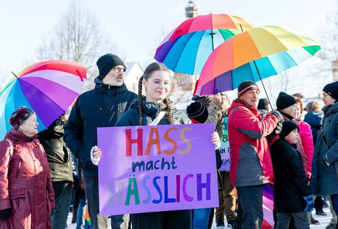 Ein Mädchen hält ein Schild mit der Aufschrift "Hass macht hässlich" in die Kamera. Im Hintergrund halten Menschen bunte Regenschirme in der Hand.