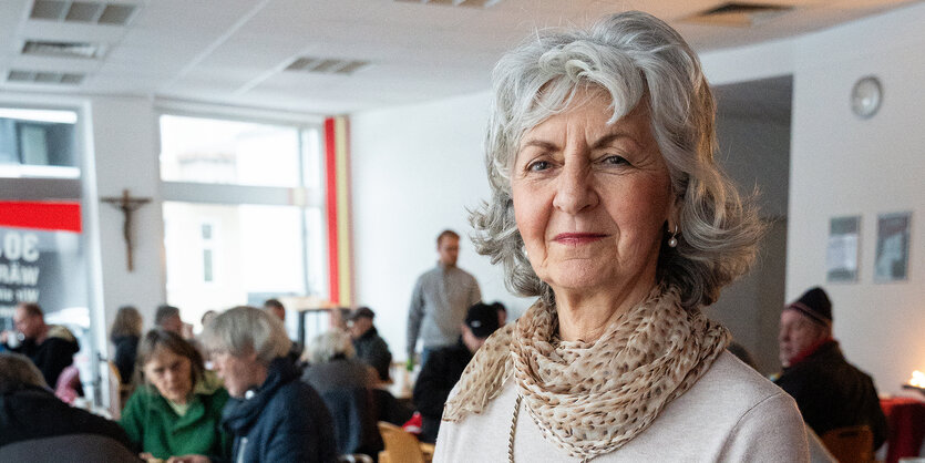 Eine ältere Dame mit grauen Locken steht in der Wärmestube. Im Hintergrund sind die Besucher:innen zu sehen.