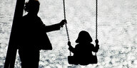Schattenriss - Frau mit Kind auf dem Spielplatz, das Kind schaukelt, die Mutter gibt der Schaukel einen Schubs