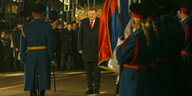 Milorad Dodik steht neben einer Flagge, vor ihm ein Soldaten in Uniform mit Flaggen, im Hintergrund stehen vor allem Männer und schauen zu