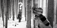 Szene aus einem Schwarz-Weiß-Film. In einem Wald liegt Schnee. Eine Person mit einer Fellmütze steht mit dem Rücken zur Kamera im Bild und schaut zwischen Baumstämmen hindurch in Richtung eines aufrecht gehenden Bibers. Der Biber ist ein:e Schauspieler:in