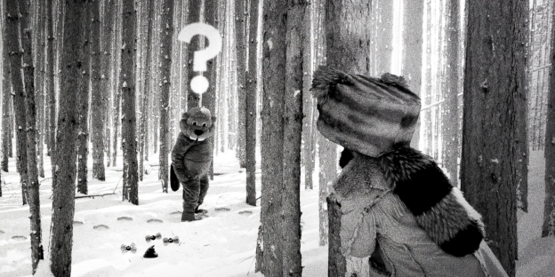 Szene aus einem Schwarz-Weiß-Film. In einem Wald liegt Schnee. Eine Person mit einer Fellmütze steht mit dem Rücken zur Kamera im Bild und schaut zwischen Baumstämmen hindurch in Richtung eines aufrecht gehenden Bibers. Der Biber ist ein:e Schauspieler:in im Biberkostüm. Über dem Biber schwebt ein weißes Fragezeichen im Bild.