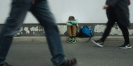 Ein Junge hockt, den Kopf auf den Knien, auf einem Fußweg, zwei Erwachsene gehen vorbei
