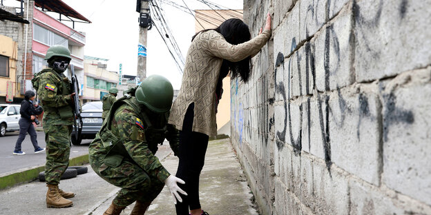 Ein Soldat tastet eine Frau ab, die ihre Hände gegen eine Mauer hält