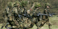 Mit Blattwerk und Gras getarnte Soldaten rennen mit ihren Waffen im Rahmen ihrer Grundausbildung bei einer Übung über das freie Gelände