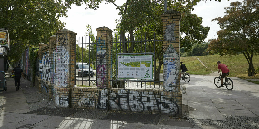 Zwei Radfahrer fahren in den Görlitzer Park, im Vordergrund ein Teil der Mauer des Parks