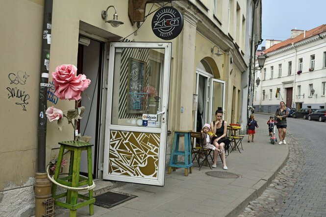 Eine Altstadtgasse, auf dem Bürgersteig sitzt eine Frau mit Kind im Straßenkaffee, eine Plastikrose auf einem Stuhl neben dem Eingang