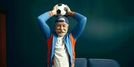 Ein älterer Mann im Trainingsanzug, mit schick onduliertem Bart hält einen Fussball über seinen Kopf