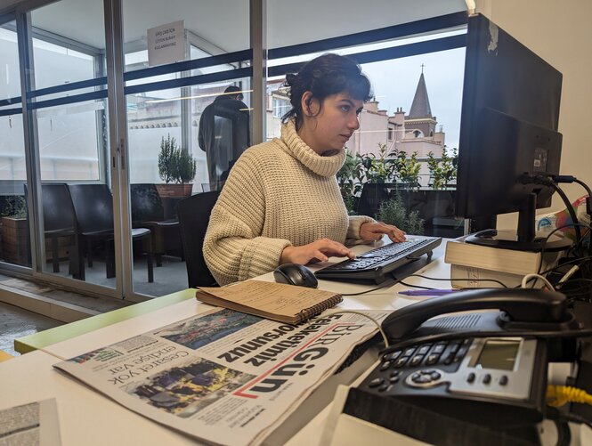 Eine Frau sitzt am Schreibtisch. Vor ihr sind Zeitungen und ein Laptop.