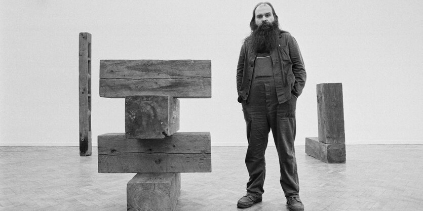 Carl Andre steht mit Bart und Arbeitshose neben einer Holzskulptur