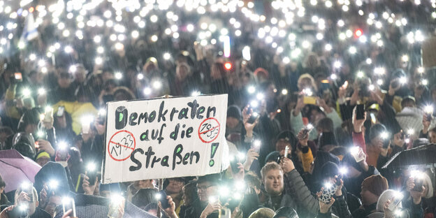 Mehrere Menschen nehmen am 23. Januar 2024 in Darmstadt an einer Demonstration gegen Rechtsextremismus und die AfD teil. Dabei steht auf einem Plakat «Demokraten auf die Straßen». Zum Abschluss der Kundgebung halten die Teilnehmer im strömenden Regen ihre Mobiltelefone in die Höhe.