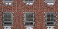 Eine Backsteinwand mit vergitterten Fenstern und Nummern daran