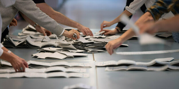 ein Tisch mit Wahlzetteln und viele Hände die sich die Zettel nehmen und auseinanderfalten