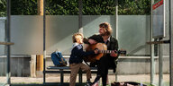 Ein Junge und ein Mann mit Gitarre in der Hand sitzen an einer Wiener Bushaltestelle