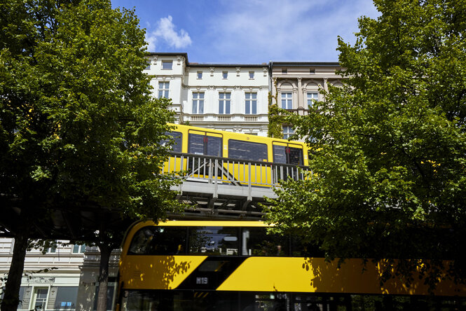 Zwei gelbe U-Bahnen kreuzen sich vor Sommerbäumen und Albauten