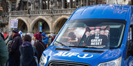 Blauer Wagen mit AUF1 Logo hat an der Frontscheibe Plakate festgesteckt mit den Köpfen von George Soros, Anthony Fauci, Bill Gates, Henry Kissinger, daneben Demonstranten vor einem Gebäude
