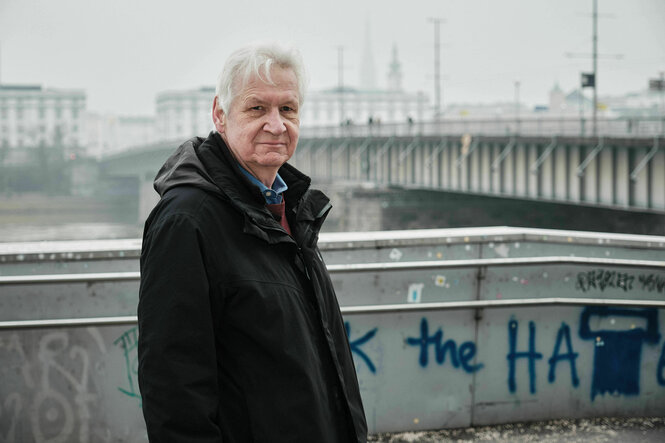 Ein alter Mann steht in dunkler Jacke vor einer Brücke . im Hintergrund Linz