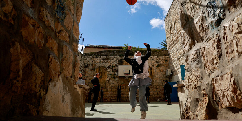 Ein Mädchen mit Kopftuch spielt auf einem ummauerten Hof Basketball, zwei Mädchen beobachten sie dabei