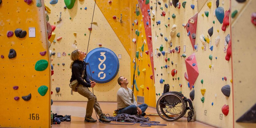 Eine Frau und ein Mann in einer Kletterhalle, sieht steht und hat ein Seil in der Hand, er sitzt neben einem Rollstuhl auf dem Boden.