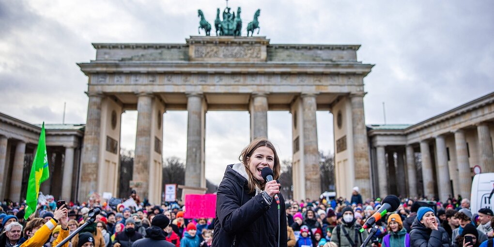 Eine Frau steht vor dem Brandenburgertor und spricht mit Mikrophon zu Demonstranten.