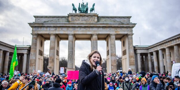 Eine Frau steht vor dem Brandenburgertor und spricht mit Mikrophon zu Demonstranten.