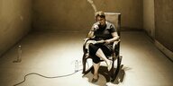 Eine Frau sitzt auf einem Stuhl in einem leeren Raum und liest aus einem Buch in ein Mikrofon rein.