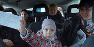 Aufnahme ins Innere eines Kleinbusses: Fünf Menschen sitzen auf den Rückbänken. In der Bildmitte sitzt ein Kind in einer rot-schwarz geblümten Winterjacke Jacke. Es trägt eine fliederfarbene Mütze und hält einen unleserlich beschrifteten Zettel in die Kamera.